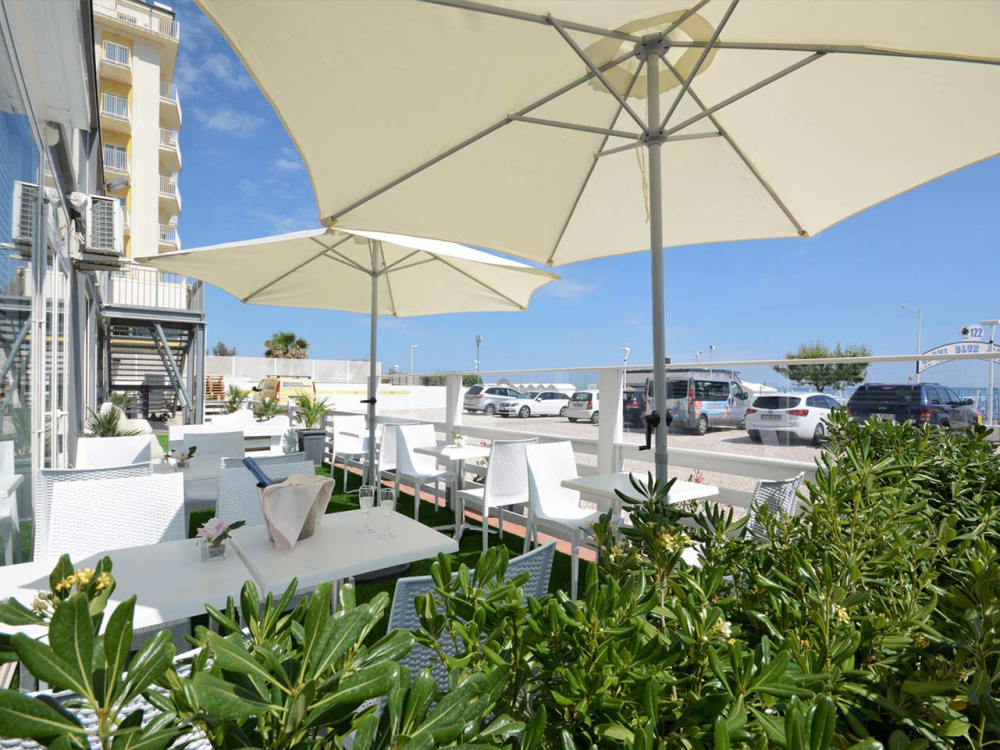 Terrazza con ombrelloni bianchi, tavoli e sedie, vista su parcheggio e mare.