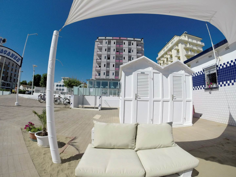 Spiaggia con cabine bianche, ombrellone e divano, edifici sullo sfondo.