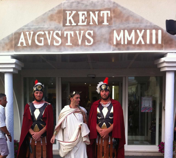 Persone in costumi romani davanti a un edificio con scritta 'KENT AVGVSTVS MMXIII'.