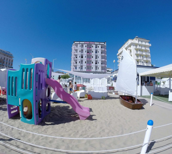 Spiaggia con giochi per bambini, barca a vela e edifici sullo sfondo.