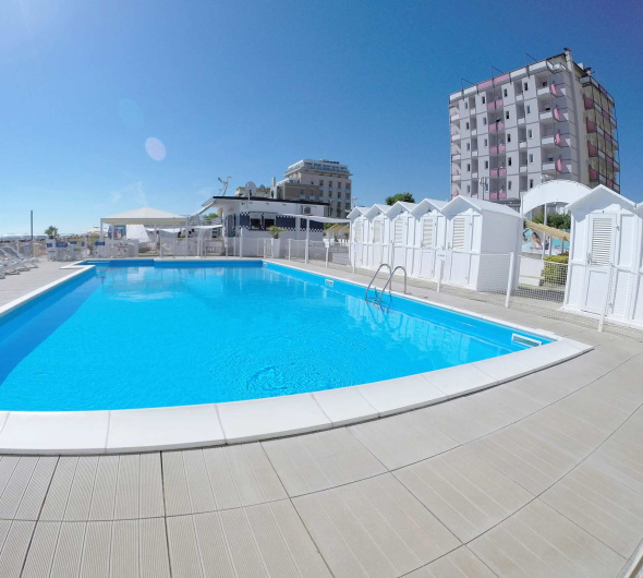 Piscina all'aperto con lettini, cabine e palazzi sullo sfondo, sotto un cielo sereno.